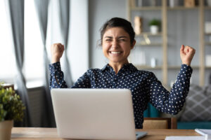 Euphoric young woman celebrate victory triumph sit at home desk with laptop computer win online fortune feel excited get new job opportunity good exam result great news concept