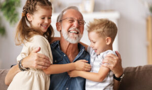 Cheerful aged man smiling and embracing cute boy and girl while resting on couch at home together