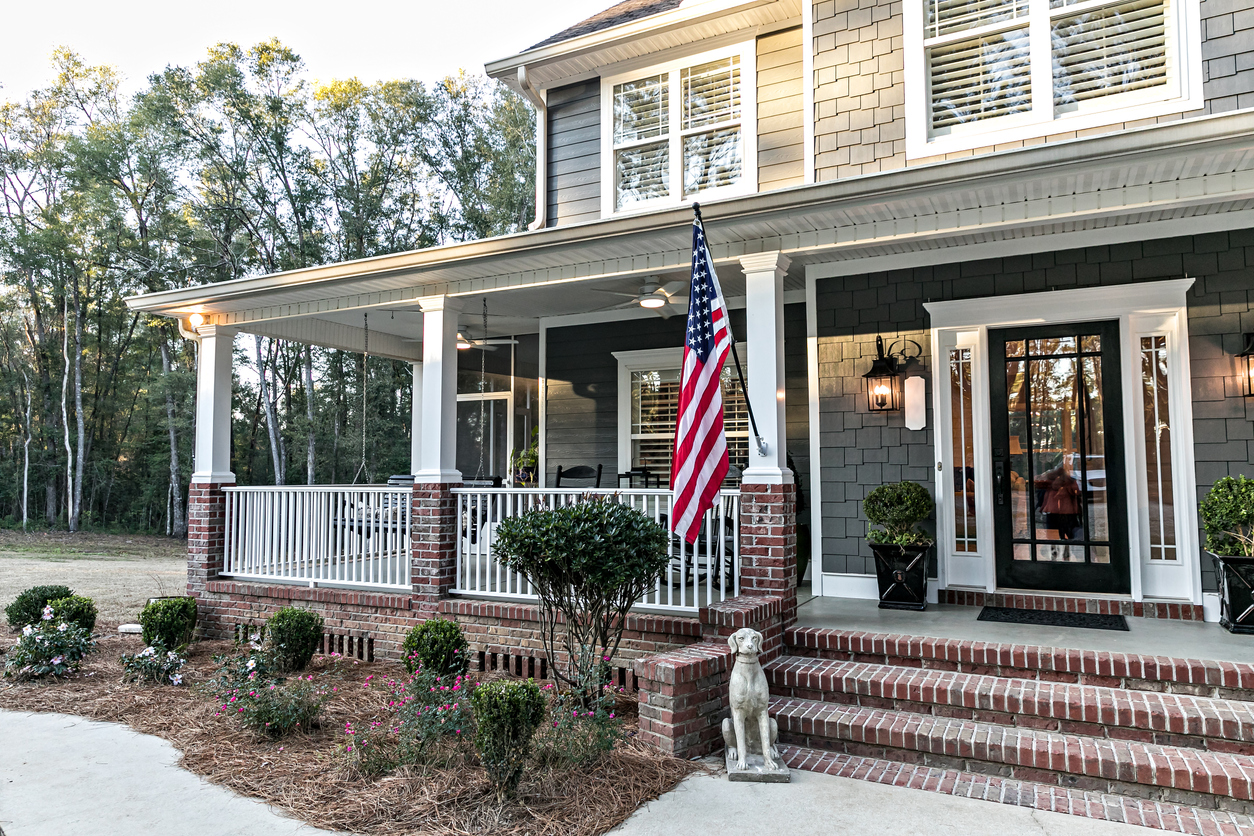 Close up of the front door entrance to a large two story blue gray house with wood and vinyl siding and a large American flag.