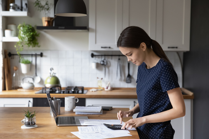 Woman use calculator calculates expenses, makes personal budget analysis, busy in planning and control of money, standing near table with bills and laptop. Savings and economy, manage finances concept